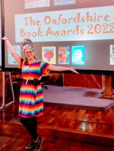 Jo Cotterill wearing a rainbow dress and a rainbow headband, standing in front of a screen showing "The Oxfordshire Book Awards 2022" and pictures of some of the winning books